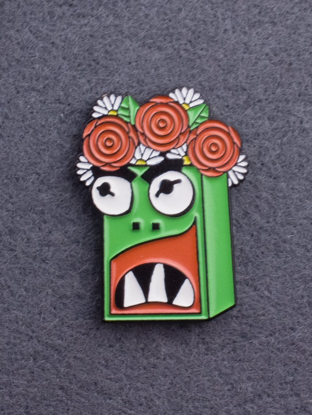 An enamel pin of Garry the kaiju. Green character with white teeth and a flower crown. 