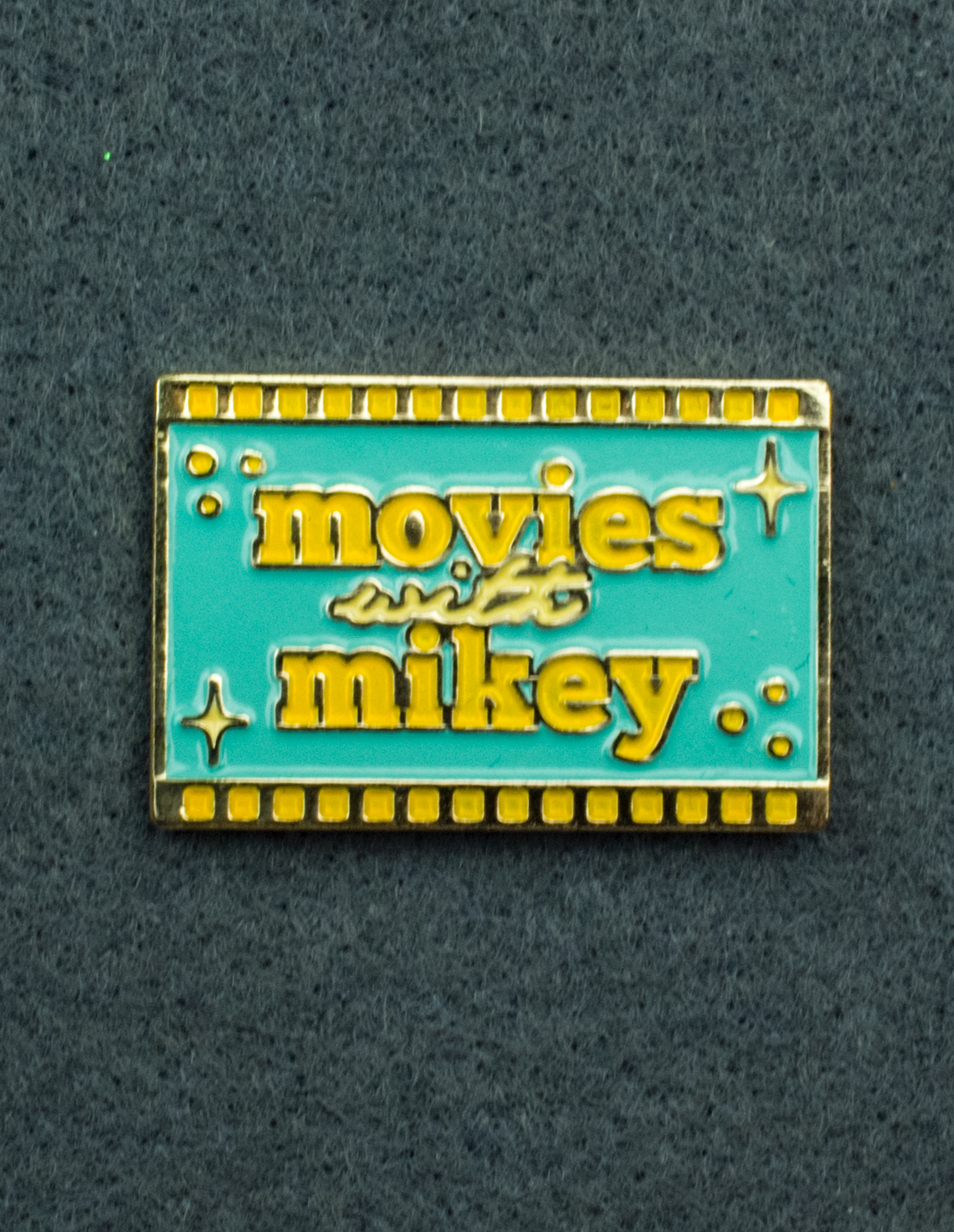 An enamel pin featuring the Movies with Mikey logo on a teal background with yellow film strips on the top and bottom. 